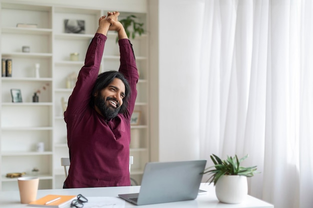 Foto homem indiano feliz esticando os braços depois de trabalhar no laptop em casa