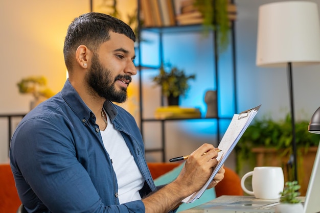 Homem indiano freelancer fazendo anotações em clipboard no escritório em casa usando laptop preparar relatório de negócios