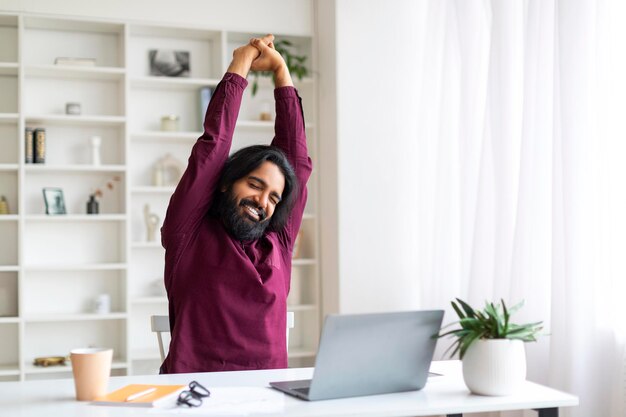 Homem indiano freelancer relaxado fazendo uma pausa e alongando-se depois de trabalhar no laptop