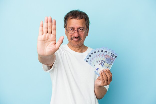 Homem indiano sênior segurando projeto de lei isolado em um fundo azul em pé com a mão estendida, mostrando o sinal de pare, impedindo-o.