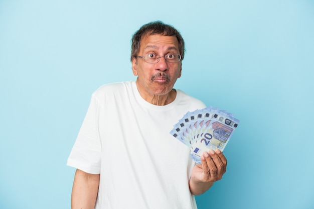 Homem indiano sênior segurando projeto de lei isolado no fundo azul encolhe os ombros e abre os olhos confusos.