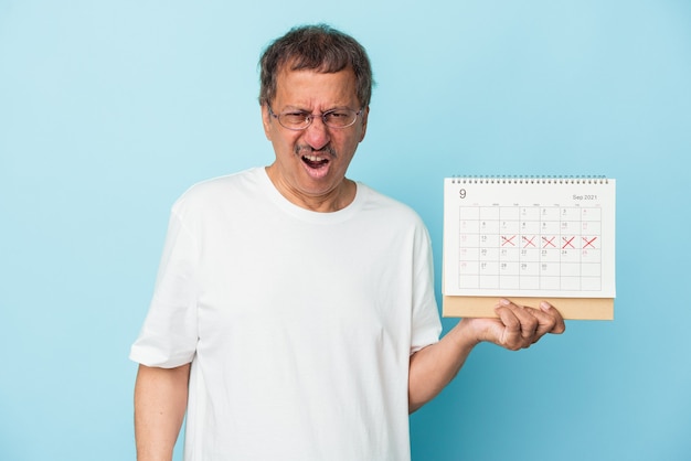 Homem indiano sênior segurando um calendário isolado no fundo azul, gritando com muita raiva e agressividade.