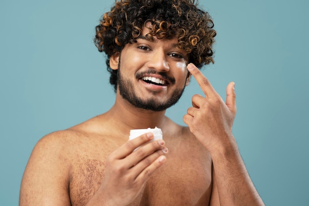 Homem indiano sorridente atraente com frasco de exploração encaracolado com creme para o corpo, olhando para a câmera. Cuidados com a pele