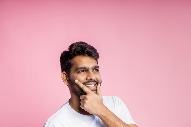 Homem indiano sorridente desvia o olhar sobre um fundo rosa, as mãos no queixo, olhar feliz pensando. camisa branca