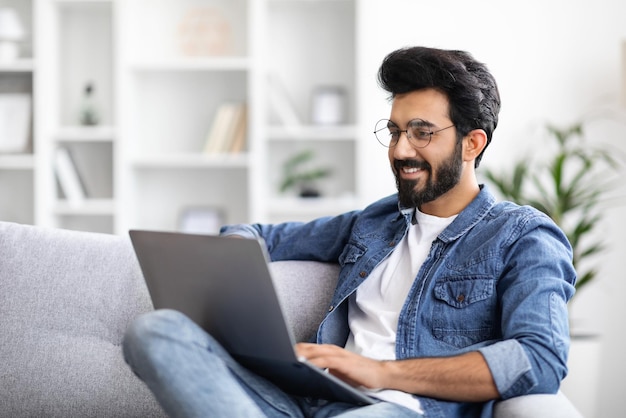 Homem indiano sorridente trabalhando em laptop enquanto está sentado no sofá em casa