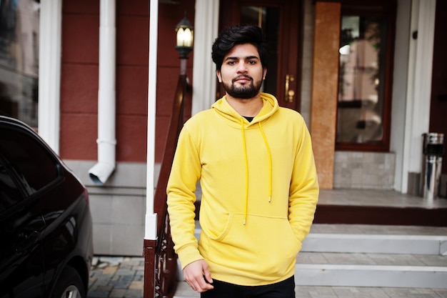 Homem indiano urbano jovem hipster em um moletom amarelo na moda cara sul-asiático legal usa capuz na varanda da casa com carro de negócios preto