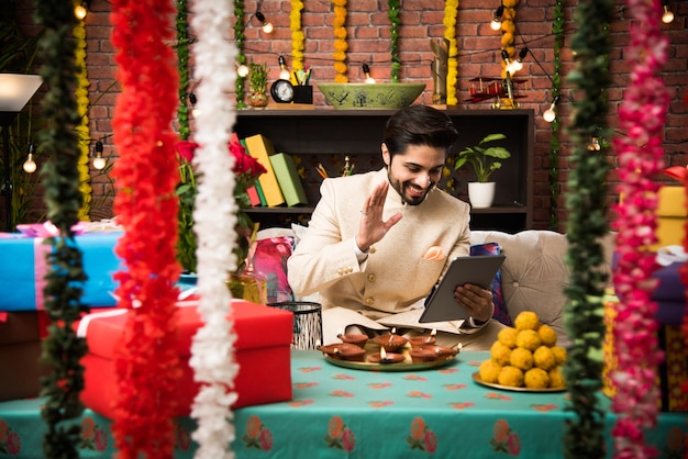 Homem indiano usando computador tablet touchscreen no dia do festival enquanto usava uma roupa tradicional. sentado no sofá com presentes e doces com iluminação bokeh