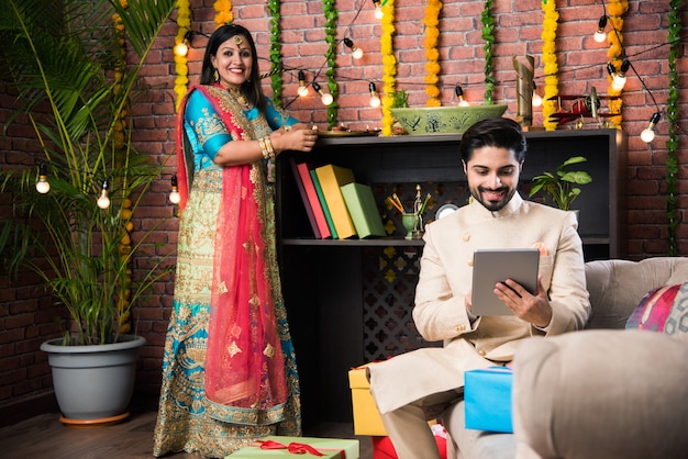 Homem indiano usando guia ou tablet touchscreen e esposa iluminando diyas ao fundo no dia do festival de Diwali