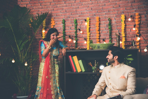 Homem indiano usando guia ou tablet touchscreen e esposa iluminando diyas ao fundo no dia do festival de Diwali