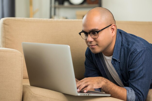 Homem indiano usando laptop no sofá na sala de mensagens de texto no laptop enviando mensagem