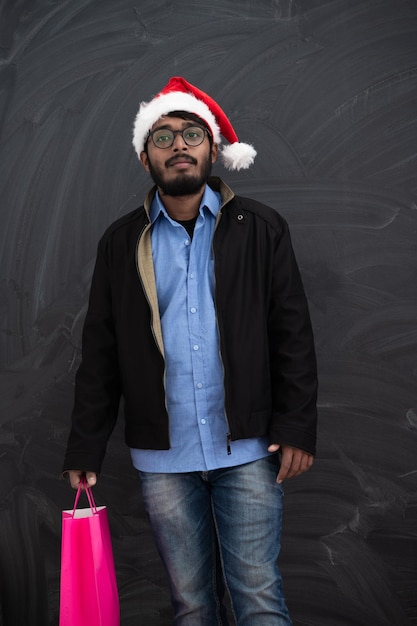 Foto homem indiano vestindo roupas de festa com chapéu de papai noel com sacola de compras no estúdio de fundo escuro pele escura do oriente médio papai noel feliz natal