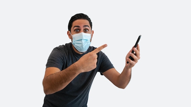 Homem indica com o dedo indicador para smartphone e máscara facial a salvo de coronavírus isolado no fundo branco. Uma pessoa mostrando um dispositivo de telefone. Conceito de quarentena pandêmica de Covid-19