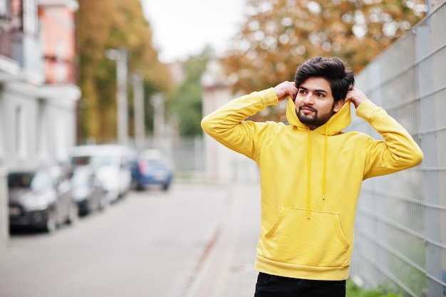 Homem índio jovem hipster urbano em um moletom amarelo na moda Cara sul-asiático legal usa capuz andando na rua de outono