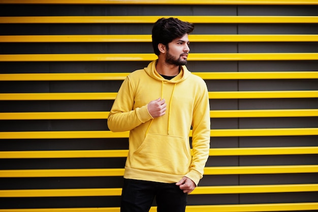 Homem índio jovem hipster urbano em um moletom amarelo na moda Cara sul-asiático legal usa capuz contra fundo listrado