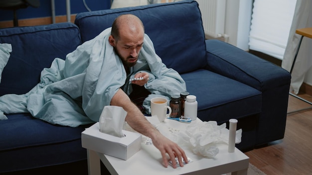 Homem indisposto à procura de comprimidos de cápsula na mesa e lendo rótulos de medicamentos contra gripe e resfriado. Adulto olhando para pílulas e drogas, tentando curar doenças com tratamento médico