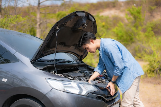 Homem infeliz novo asiático que inspeciona o motor de automóveis quebrado na frente da capa aberta carro dividido no serviço de assistência rodoviária à espera da estrada secundária.