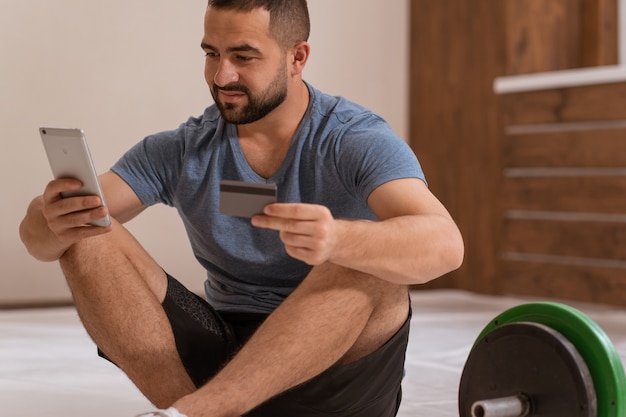 Homem instrutor de fitness com smartphone e cartão de débito ou crédito, comprando equipamentos online
