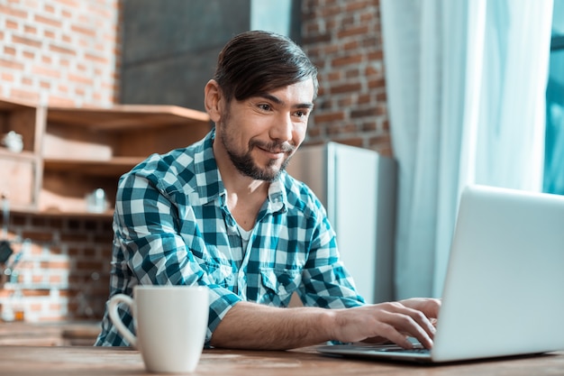 Homem inteligente positivo e alegre sentado em frente ao computador e sorrindo enquanto trabalha remotamente de casa