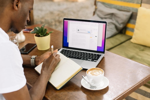 Homem interracial retrovisor usando laptop escrevendo notas no livro e tomando café no café com tecnologia sem fio