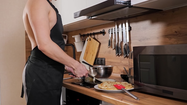 Homem irreconhecível com um torso nu e um avental preto prepara panquecas na cozinha.