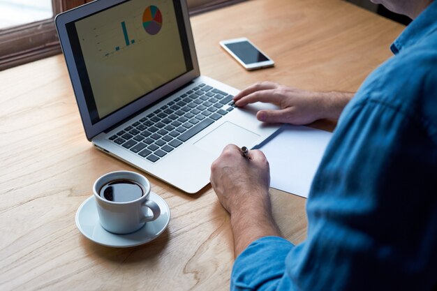 Homem irreconhecível escreve plano no papel e olha para gráficos na tela do computador, laptop.