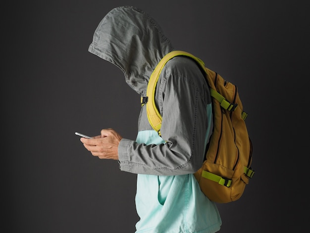 Foto homem irreconhecível no moletom com capuz e mochila amarela usando smartphone.