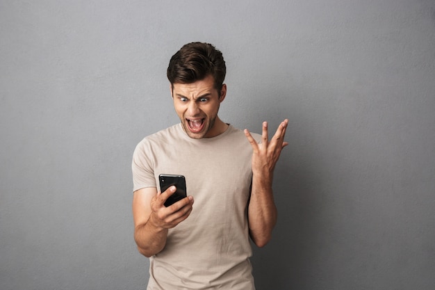 Homem irritado com cabelo escuro curto, gritando de raiva enquanto segura o smartphone