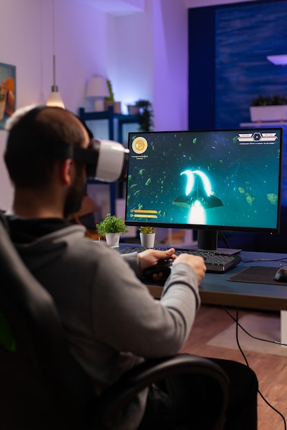 Homem jogador jogando videogame no computador poderoso tarde da noite usando fone de ouvido vr. Jogador empolgado usando controlador sem fio para atirador de espaço de jogo em torneio virtual em casa
