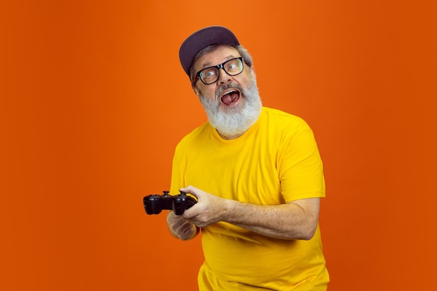 Homem jogador. Retrato de homem hippie sênior usando dispositivos, gadgets isolados em fundo laranja do estúdio. Tecnologia e conceito de estilo de vida idoso alegre. Cores da moda, juventude para sempre. Copyspace para seu anúncio.