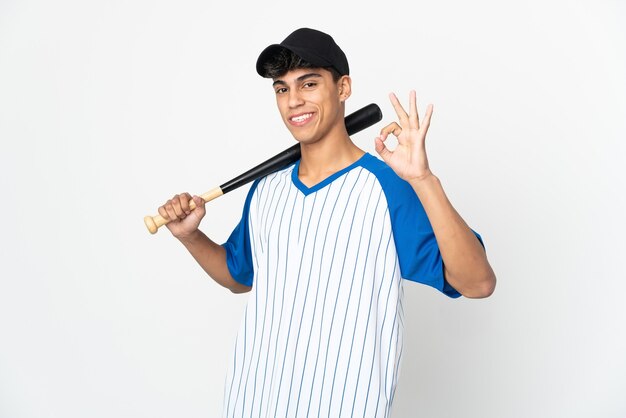 Homem jogando beisebol na parede branca isolada, mostrando um sinal de ok com os dedos