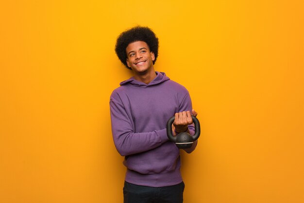 Homem jovem afro-americano fitness segurando um haltere sorrindo confiante e cruzando os braços, olhando para cima