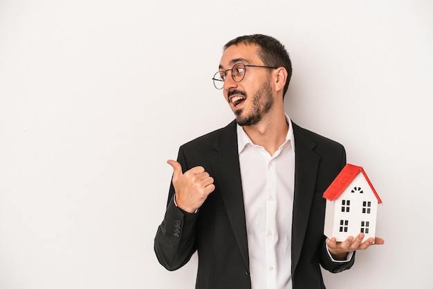 Homem jovem agente imobiliário segurando uma casa modelo isolada no fundo branco aponta com o dedo polegar afastado, rindo e despreocupado.