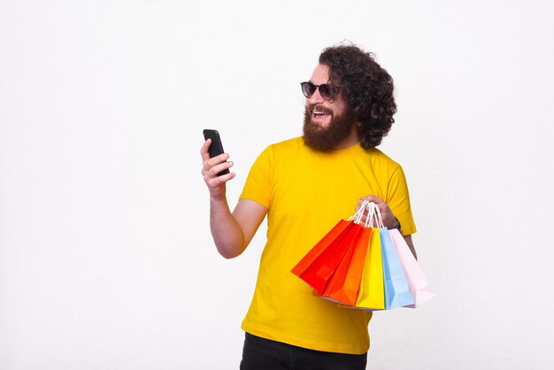 Homem jovem alegre e hippie com camiseta amarela, pagando online e segurando sacolas de compras