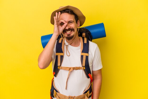 Homem jovem alpinista caucasiano isolado em fundo amarelo animado, mantendo o gesto ok no olho.