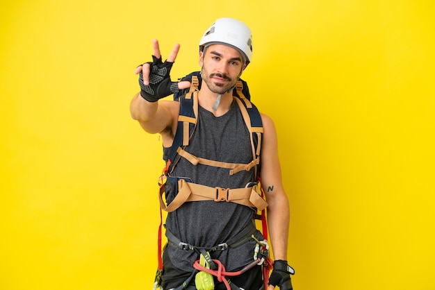 Homem jovem alpinista caucasiano isolado em fundo amarelo feliz e contando três com os dedos