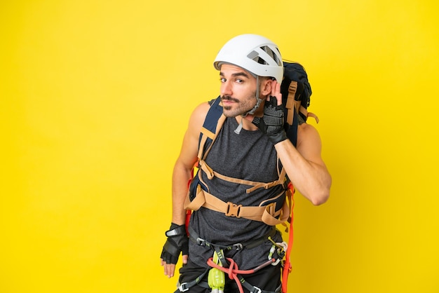 Homem jovem alpinista caucasiano isolado em fundo amarelo ouvindo algo colocando a mão na orelha