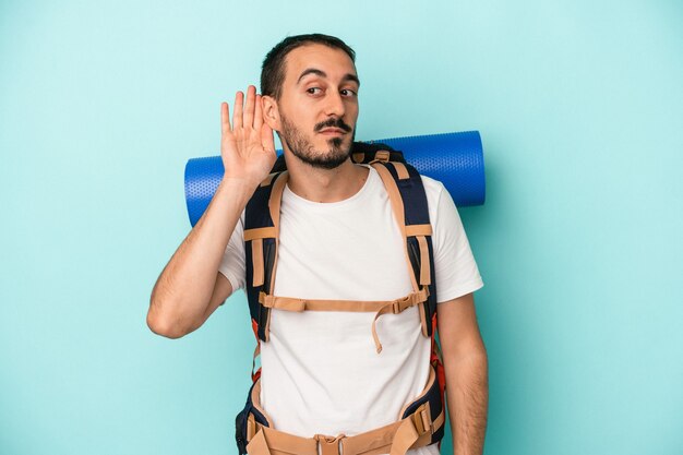 Homem jovem alpinista caucasiano isolado sobre fundo azul, tentando ouvir uma fofoca.