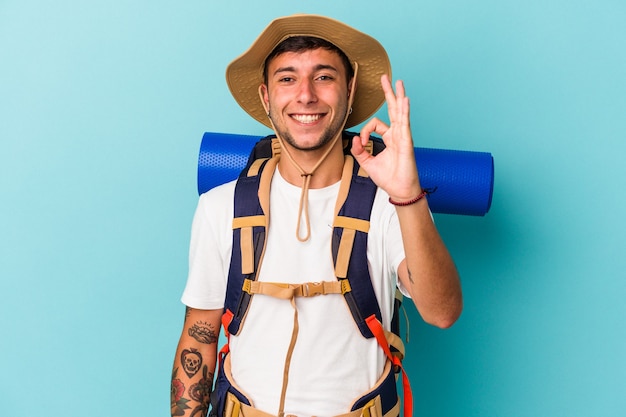 Homem jovem alpinista com chapéu isolado em fundo azul alegre e confiante, mostrando o gesto de ok.