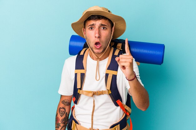 Homem jovem alpinista com chapéu isolado em um fundo azul, tendo uma ideia, o conceito de inspiração.