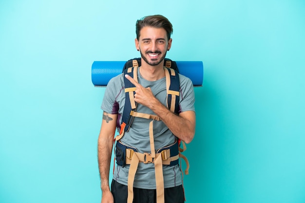 Homem jovem alpinista com uma mochila grande isolada em fundo azul, apontando para o lado para apresentar um produto