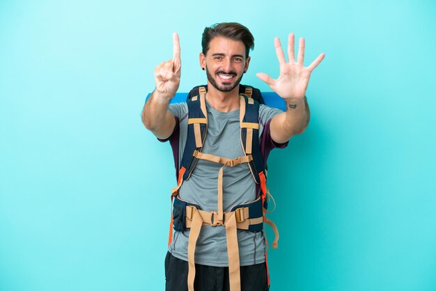 Homem jovem alpinista com uma mochila grande isolada em fundo azul, contando seis com os dedos