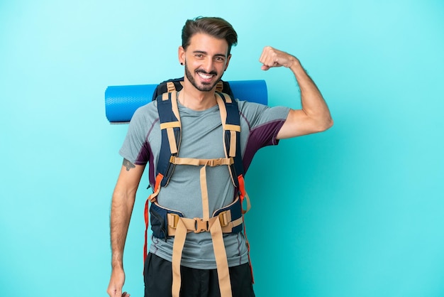 Homem jovem alpinista com uma mochila grande isolada em fundo azul, fazendo um gesto forte