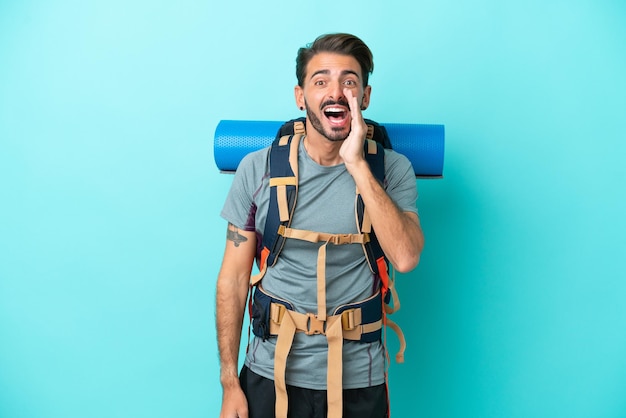 Homem jovem alpinista com uma mochila grande isolada em fundo azul gritando com a boca aberta