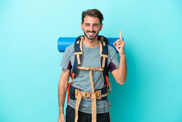 Homem jovem alpinista com uma mochila grande isolada em fundo azul mostrando e levantando um dedo em sinal do melhor