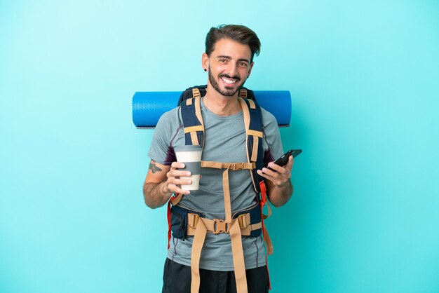 Homem jovem alpinista com uma mochila grande isolada em fundo azul segurando café para levar e um celular