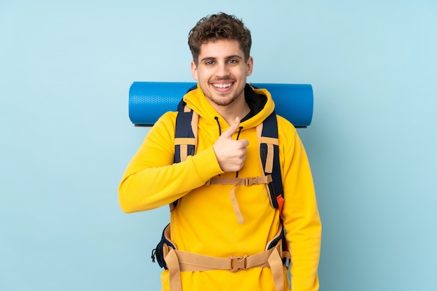 Homem jovem alpinista com uma mochila grande isolada na parede azul, dando um polegar para cima gesto