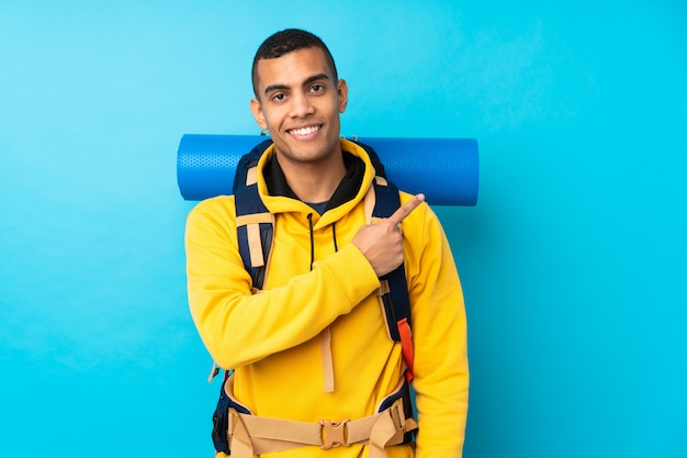 Homem jovem alpinista com uma mochila grande sobre parede azul isolada, apontando o dedo para o lado