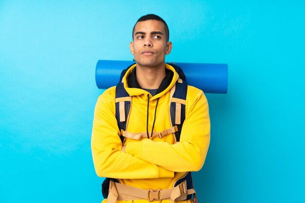 Homem jovem alpinista com uma mochila grande sobre parede azul isolada, pensando uma idéia