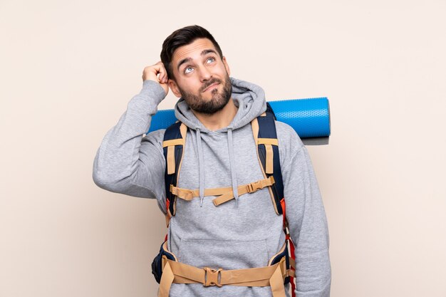 Homem jovem alpinista com uma mochila grande sobre parede isolada, tendo dúvidas e com a expressão do rosto confuso