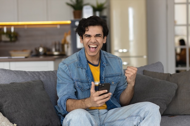 Homem jovem animado com smartphone, sente-se no sofá em casa, lendo uma boa notícia inesperada no celular. Homem feliz surpreso com a vitória na loteria online